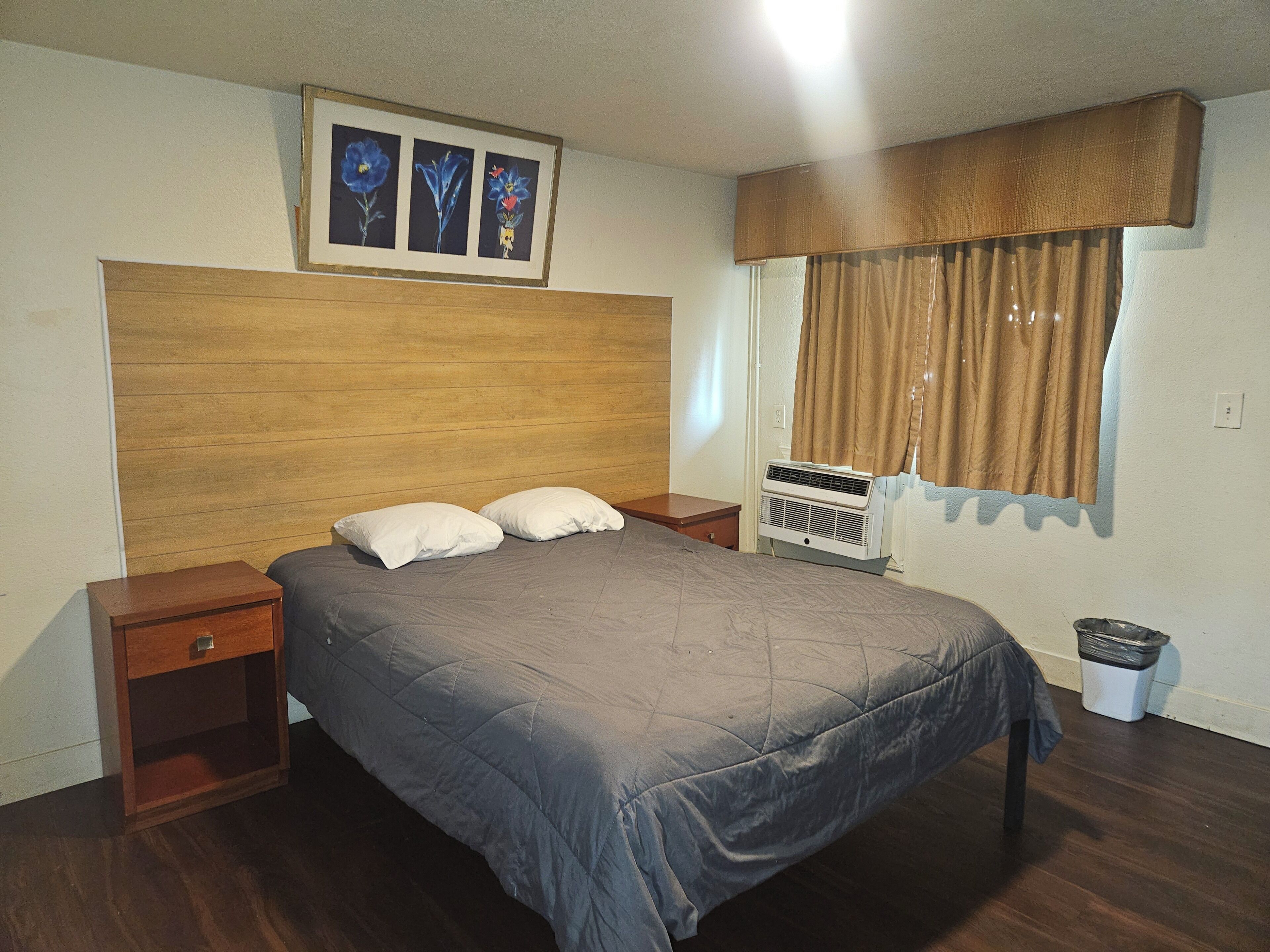 Un dormitorio con cabecera de madera, cama con ropa de cama gris, dos mesitas de noche, aire acondicionado instalado en la pared, una ventana con cortinas e imagen enmarcada sobre la cabecera.