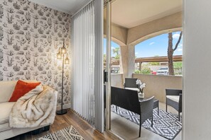 Ein Zimmer mit einer Couch, einer Wand mit Muster, einer Stehlampe, einer Schiebetür zu einem Außenbereich mit Sitzgelegenheiten, Stühlen und einem Tisch.