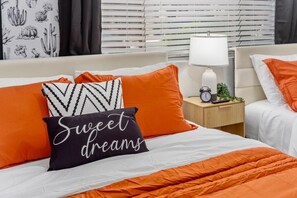 Habitación de hotel con cama, ropa de cama naranja, mesita de noche con lámpara y ventana con persianas.