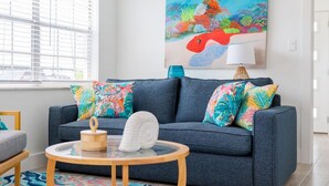 A living room with a blue sofa, colorful pillows, a round wooden table, and a painting of a fish on the wall. - Colorful Coastal Retreat Near Honeymoon Island Beaches (Dunedin)