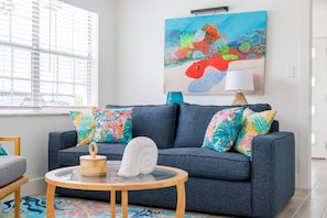 A living room with a blue sofa, colorful pillows, a round wooden table, and a painting of a fish on the wall. - Colorful Coastal Retreat Near Honeymoon Island Beaches (Dunedin)