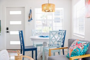 A dining area with a round table, blue chairs, a hanging light fixture, and a window with blinds. - Colorful Coastal Retreat Near Honeymoon Island Beaches (Dunedin)