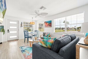 A modern living room with a grey sofa, colorful pillows, a dining area with blue chairs, and large windows with blinds. - Colorful Coastal Retreat Near Honeymoon Island Beaches (Dunedin)