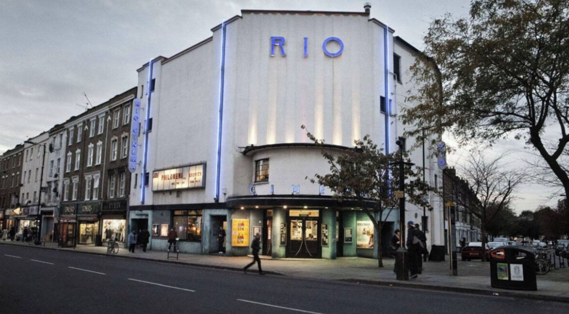 Ein Kino mit der Aufschrift 'RIO' an der Fassade, gelegen in einer Straße mit Fußgängern und parkenden Autos.