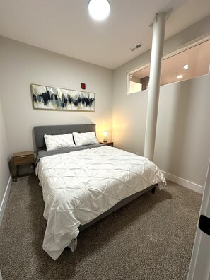 Quarto com cama, mesas de cabeceira, luminária na parede, abertura no teto e um quadro.