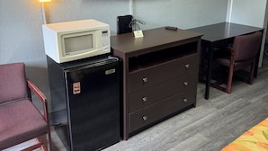 A hotel room with a flat-screen TV, a microwave, a refrigerator, a desk with drawers, a chair, and a bed with a colorful blanket.