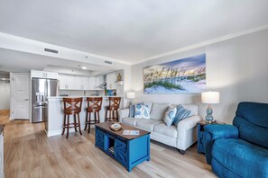 Ein modernes Wohnzimmer mit blauem Couchtisch, einem weißen Sofa mit gemusterten Kissen, einem blauen Sessel und einer Küchenzeile mit weißen Schränken und einem Edelstahlkühlschrank.