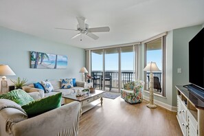 Ein Wohnzimmer mit einer Couch, Sesseln, einem Couchtisch und einem Fernseher. Es gibt einen Balkon mit Blick auf den Strand.