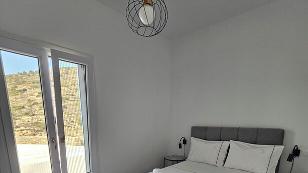 A bedroom with a large bed, white linens, a grey headboard, a bedside table with a lamp, and a view through a sliding door. - Alkinoi Residences (Sifnos)