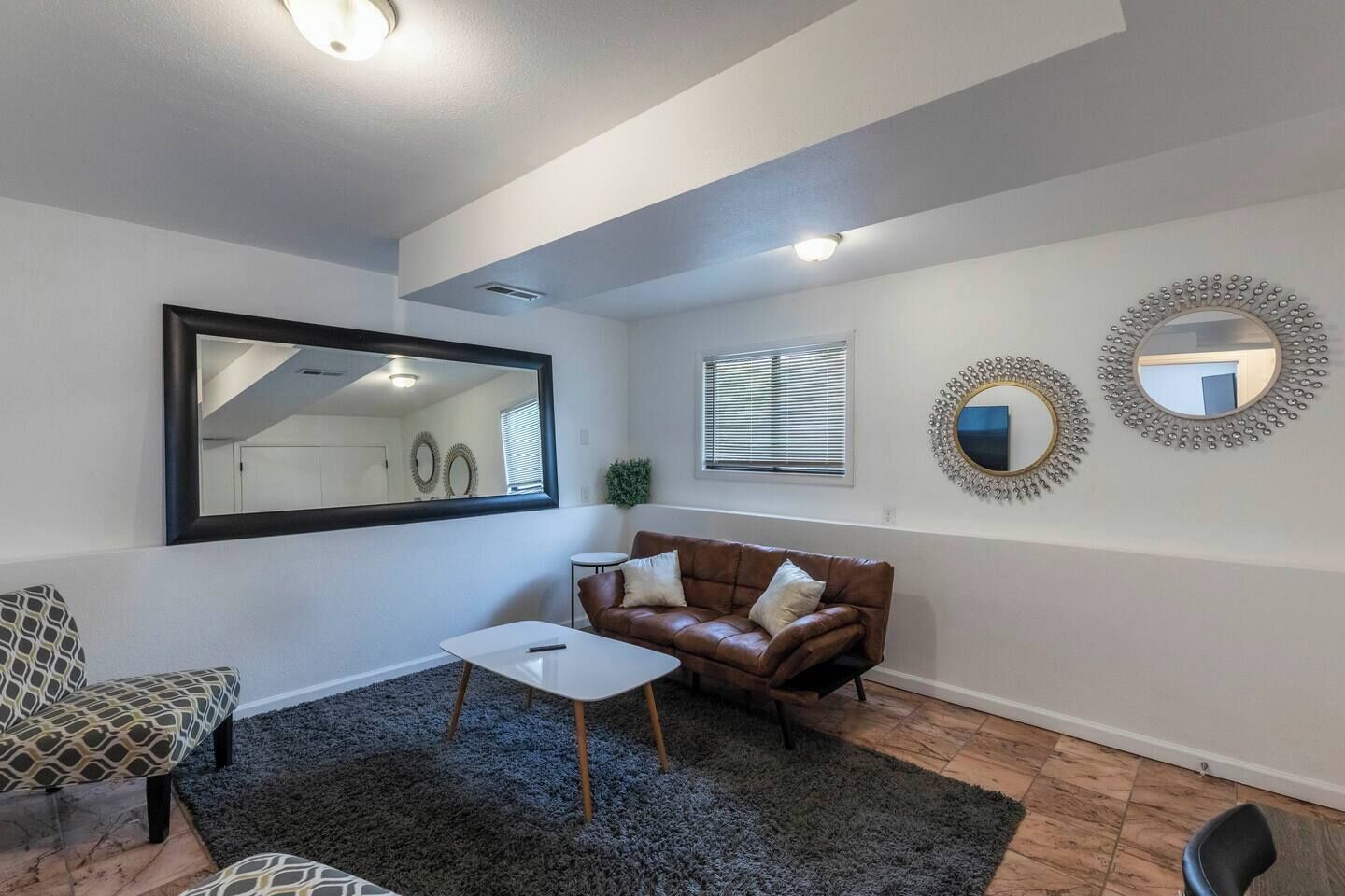 Een woonkamer met een bruine leren bank, een witte salontafel, een fauteuil met een patroon en een grote spiegel aan de muur.