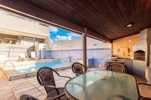 Ein Poolbereich mit Glastisch und Stühlen, einem Backofen aus Ziegeln und Blick auf ein Gebäude mit einer Kachelwand.
