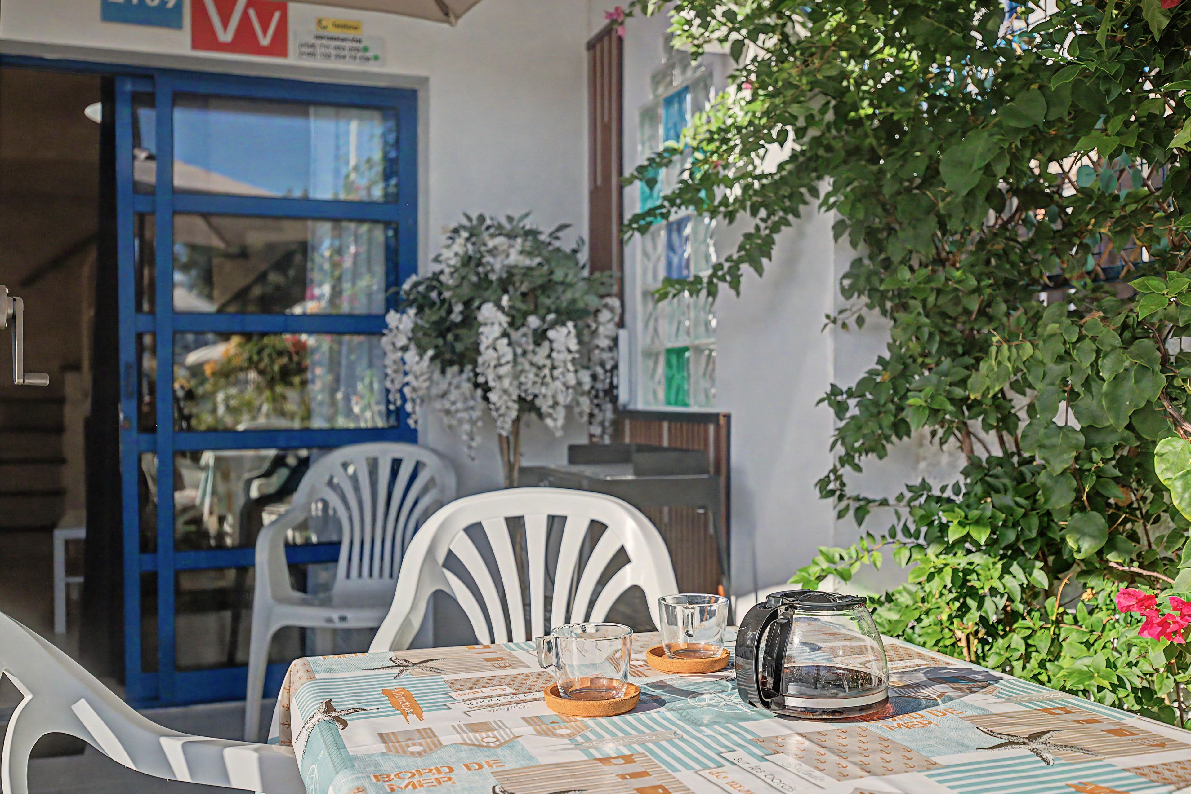 Un área de estar al aire libre con una mesa dispuesta para dos personas, una cafetera y una jarra de vidrio.