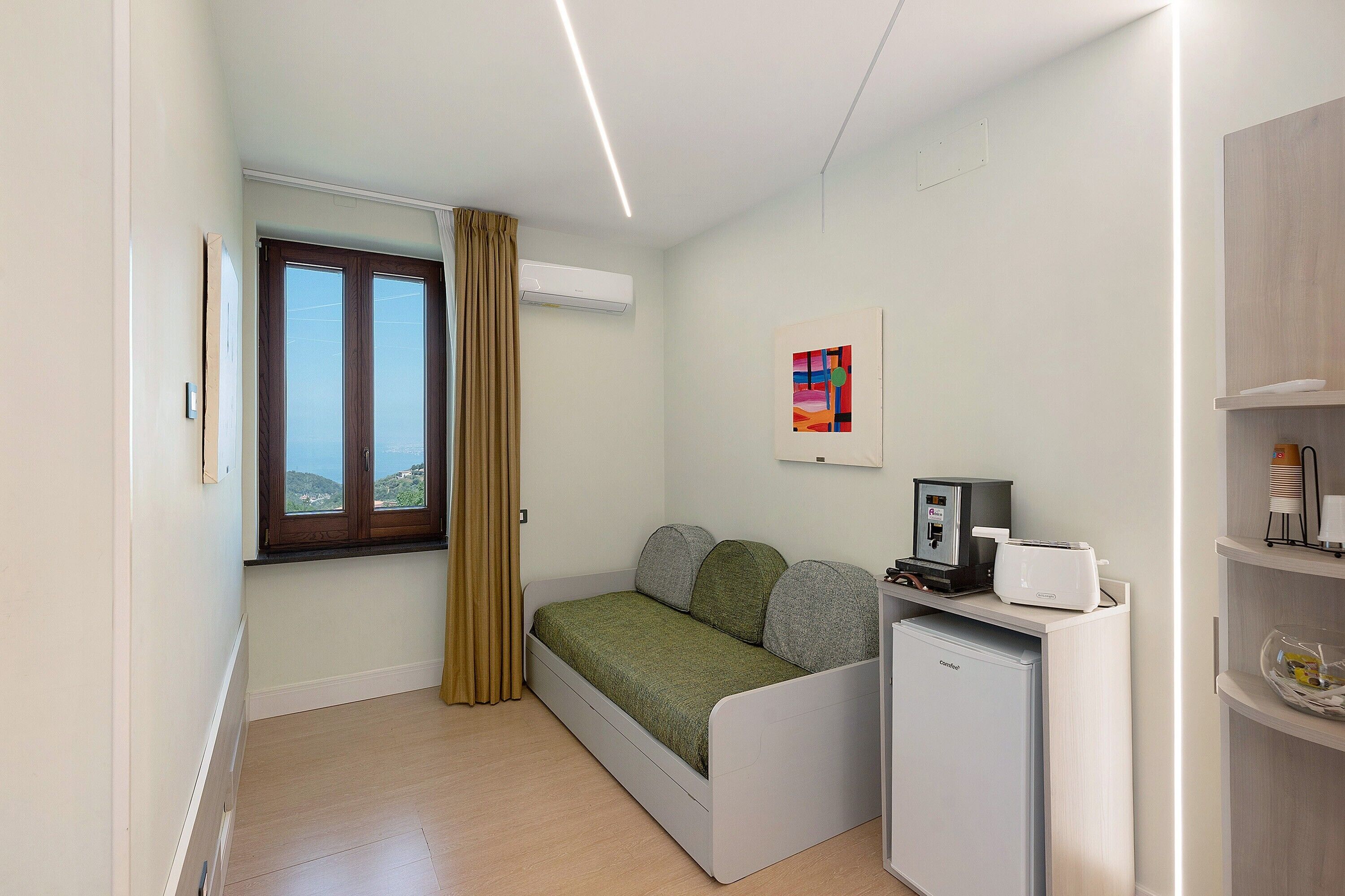 A small, compact room with a bed, a desk with a coffee machine, an air conditioner, and a window with a view.