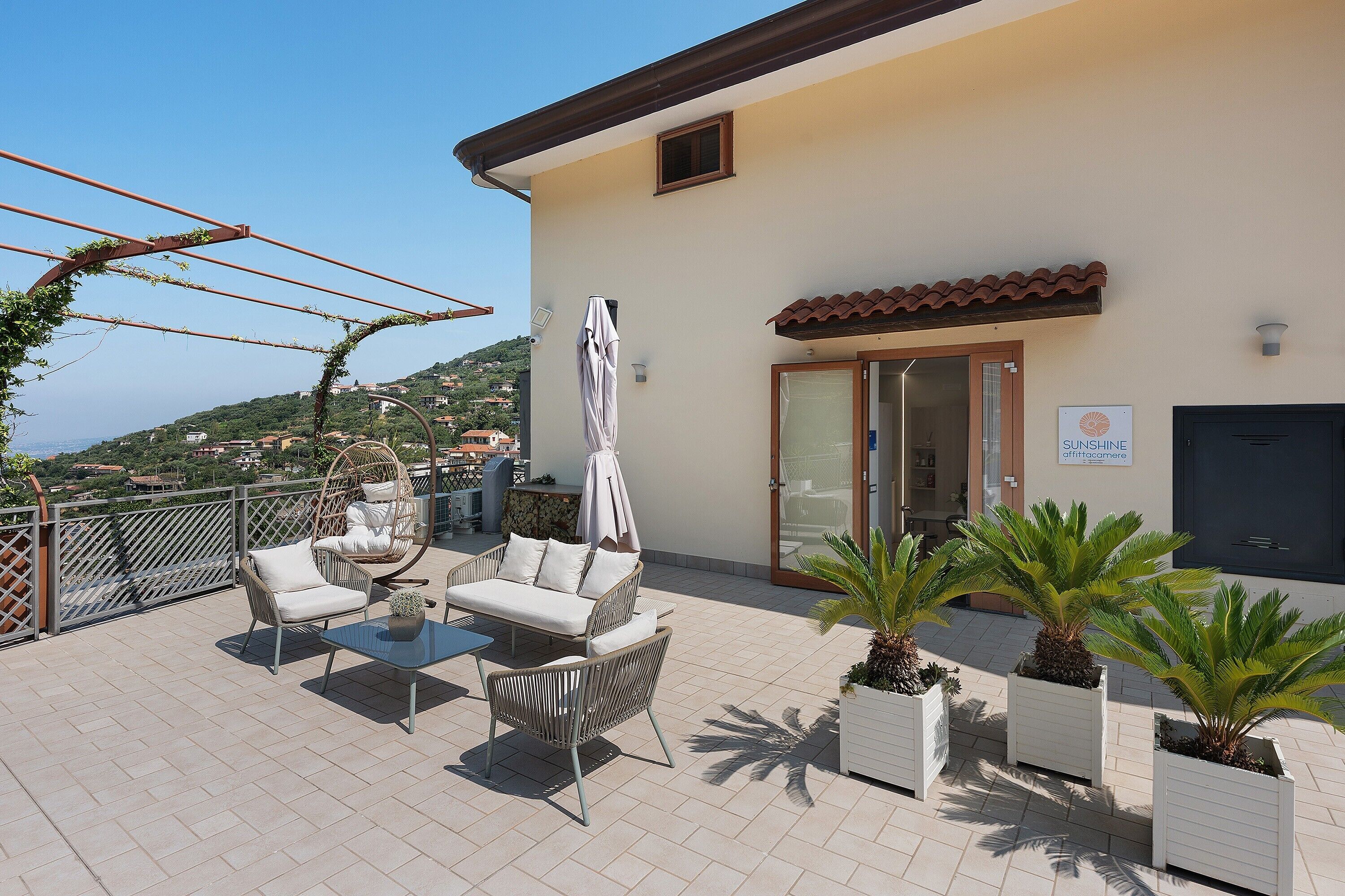 A rooftop terrace with outdoor furniture, a pergola with vines, and a view of the surrounding landscape.