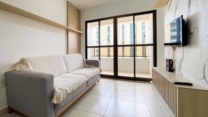 Ein Wohnzimmer mit grauem Sofa, einem an der Wand befestigten Fernseher und einem Fenster mit Blick auf Gebäude.