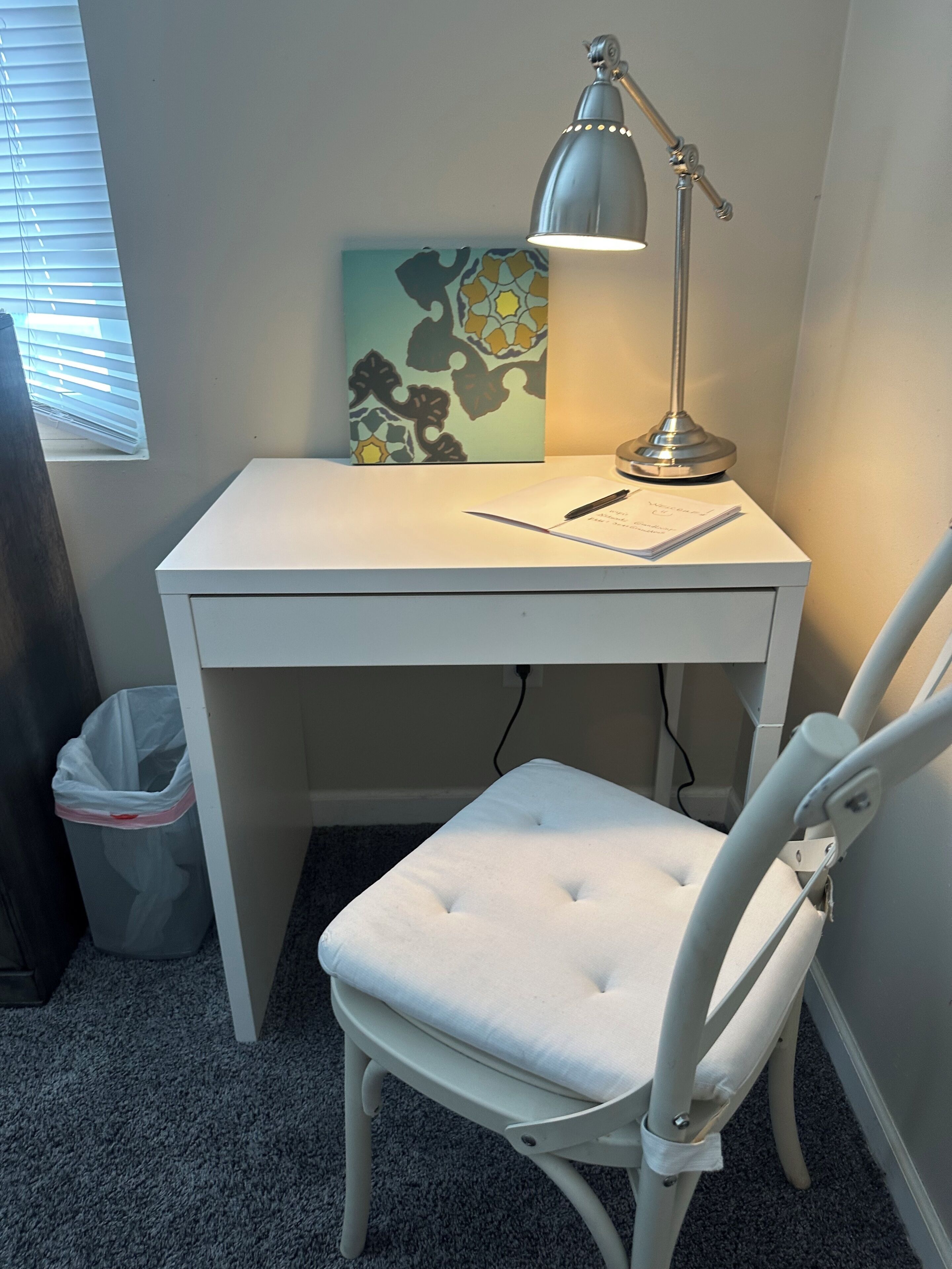 Een wit bureau met een lamp, een stoel, een prullenbak en een raam met jaloezieën.