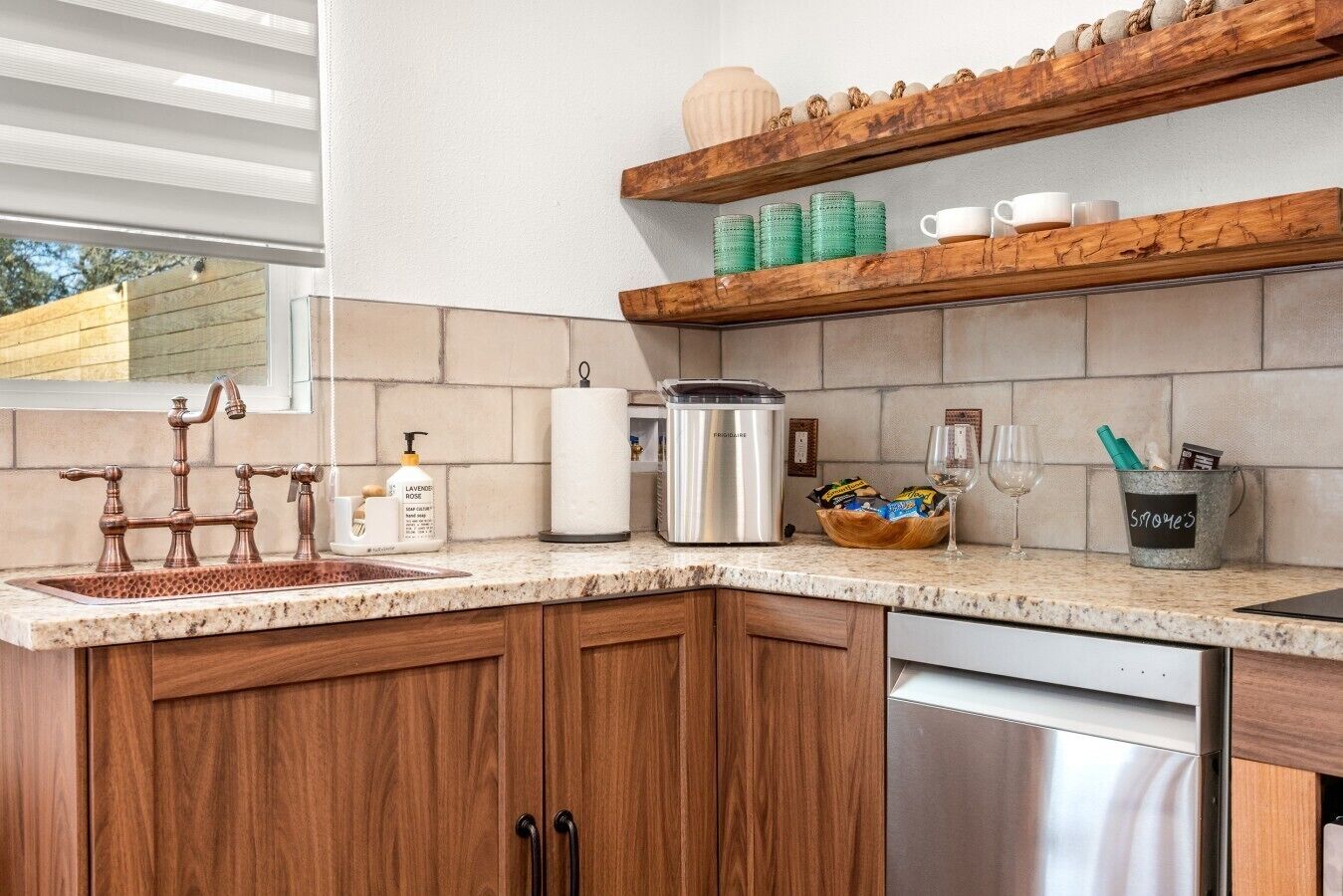 A kitchen with a copper faucet, a dish soap dispenser, a paper towel holder, a stainless steel kettle, a basket of snacks, and a mug holder with a label.