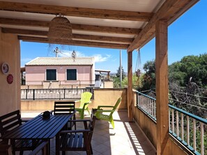 Ein überdachter Außenbereich mit Holztisch und Stühlen, einer hängenden Leuchte und Blick auf ein Gebäude mit Ziegeldach.