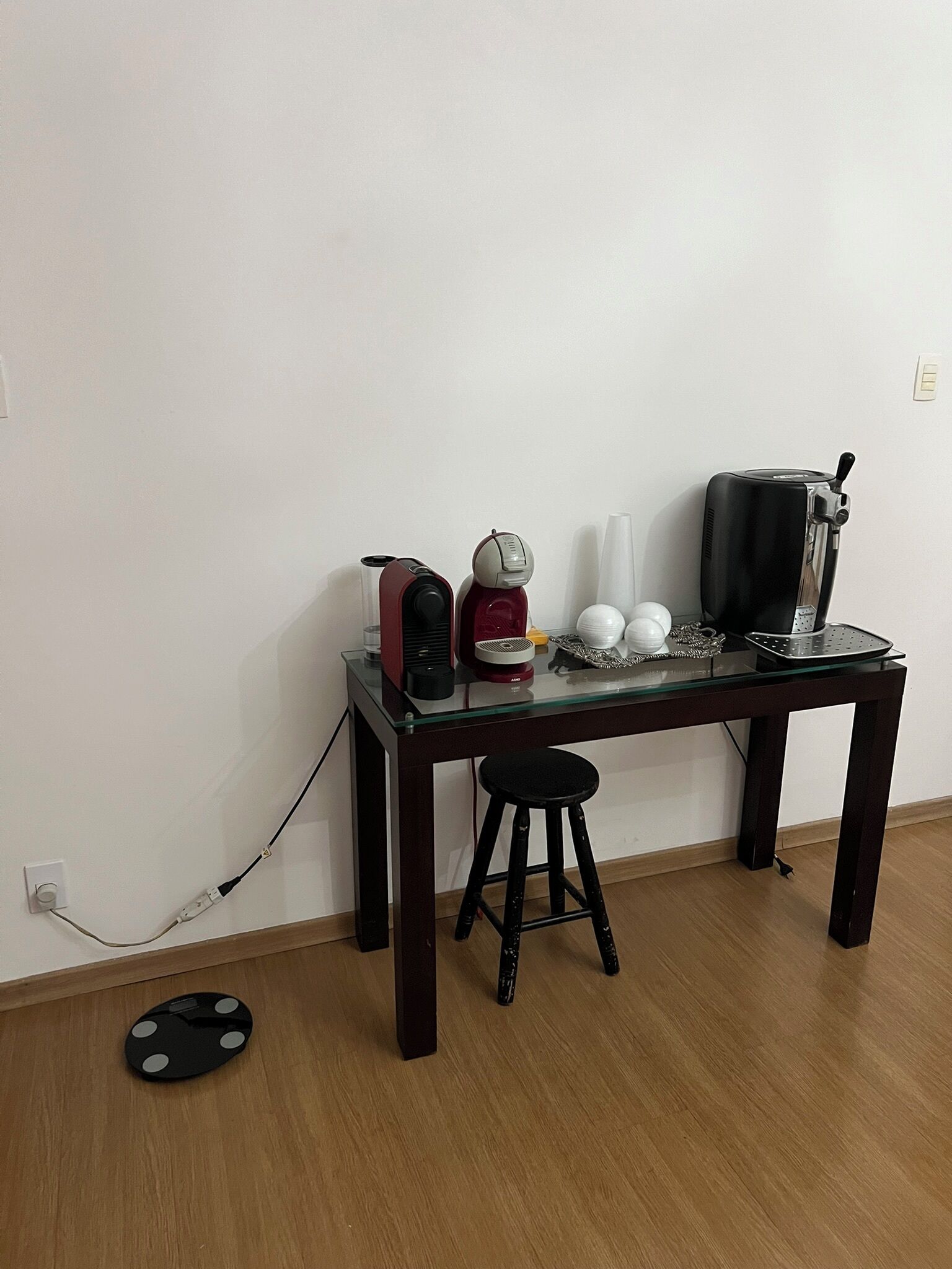 Ett litet, mörkt träbord med en glasskiva, en svart pall och en kaffemaskin.