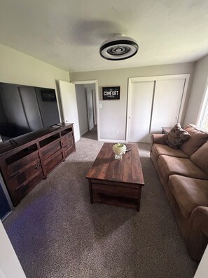 A living room with a flat-screen TV, a wooden coffee table with a vase, a brown leather sofa, and a sign on the wall that reads 'COMFORT'.