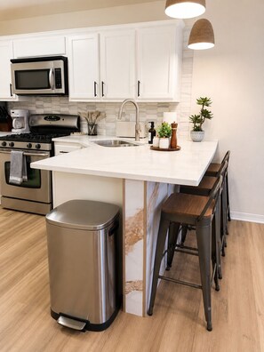 Una cocina moderna con muebles blancos, electrodomésticos de acero inoxidable, una isla central con taburetes y un bote de basura.