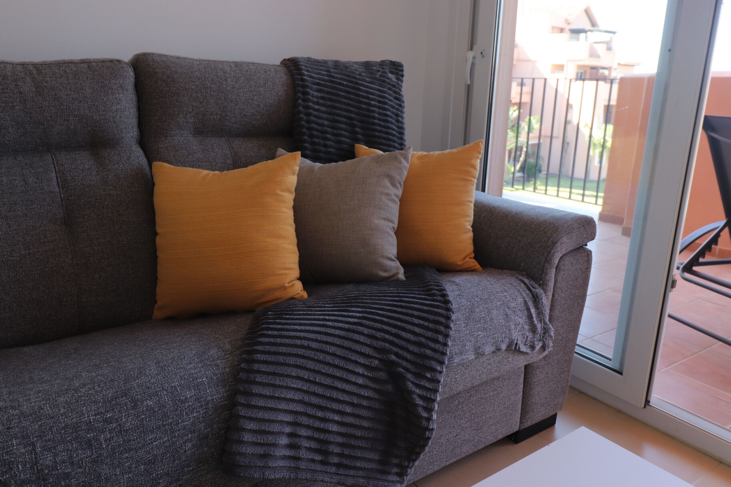 A grey sofa with three cushions in different shades, a grey throw blanket, and a sliding glass door leading to an outdoor balcony.