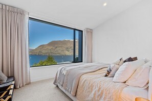 Ein Schlafzimmer mit großem Fenster, einem Bett mit vielen Kissen und Blick auf einen See und Berge.