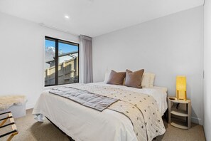 A bedroom with a large bed, a bedside table with a lamp, a window with a view of buildings, and a fluffy chair. - Aspen Ridge by MajorDomo | Fernhill Villa with Lake Views, Infrared Sauna, Spa Pool & Outdoor Fireplace (Queenstown)