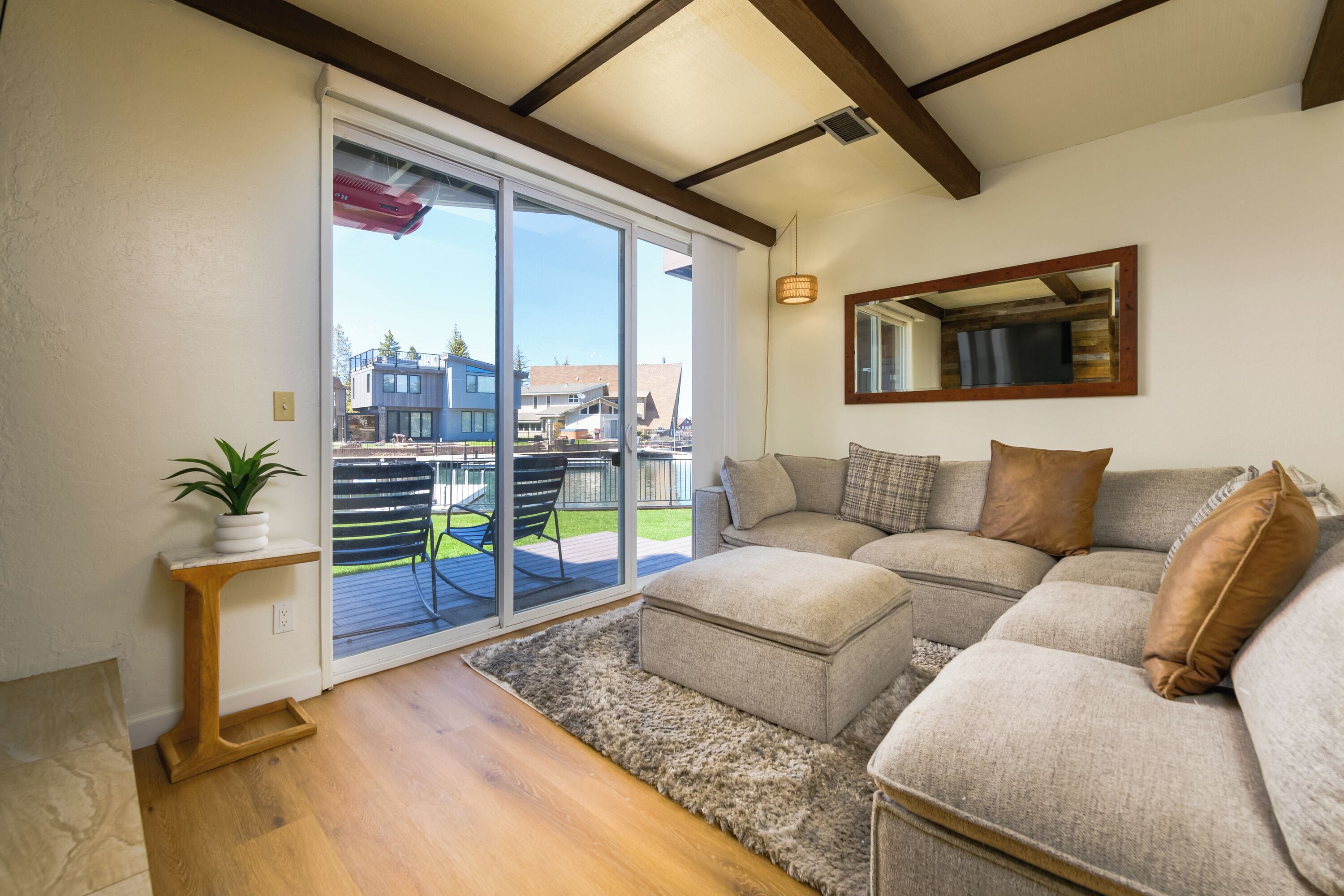 A modern living room with a sectional sofa, a wooden floor, a sliding glass door leading to a balcony, and a small side table with a plant.