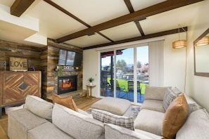 A living room with a fireplace, a television, and a view of a green landscape through large sliding doors. - Waterfront Tahoe Keys Condo. Private Boat Dock, Lake Views & Family Friendly (South Lake Tahoe)