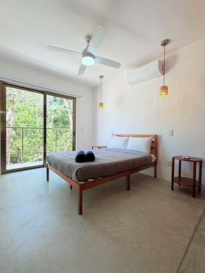Un dormitorio con cama, ventilador de techo, ventana y una mesita.