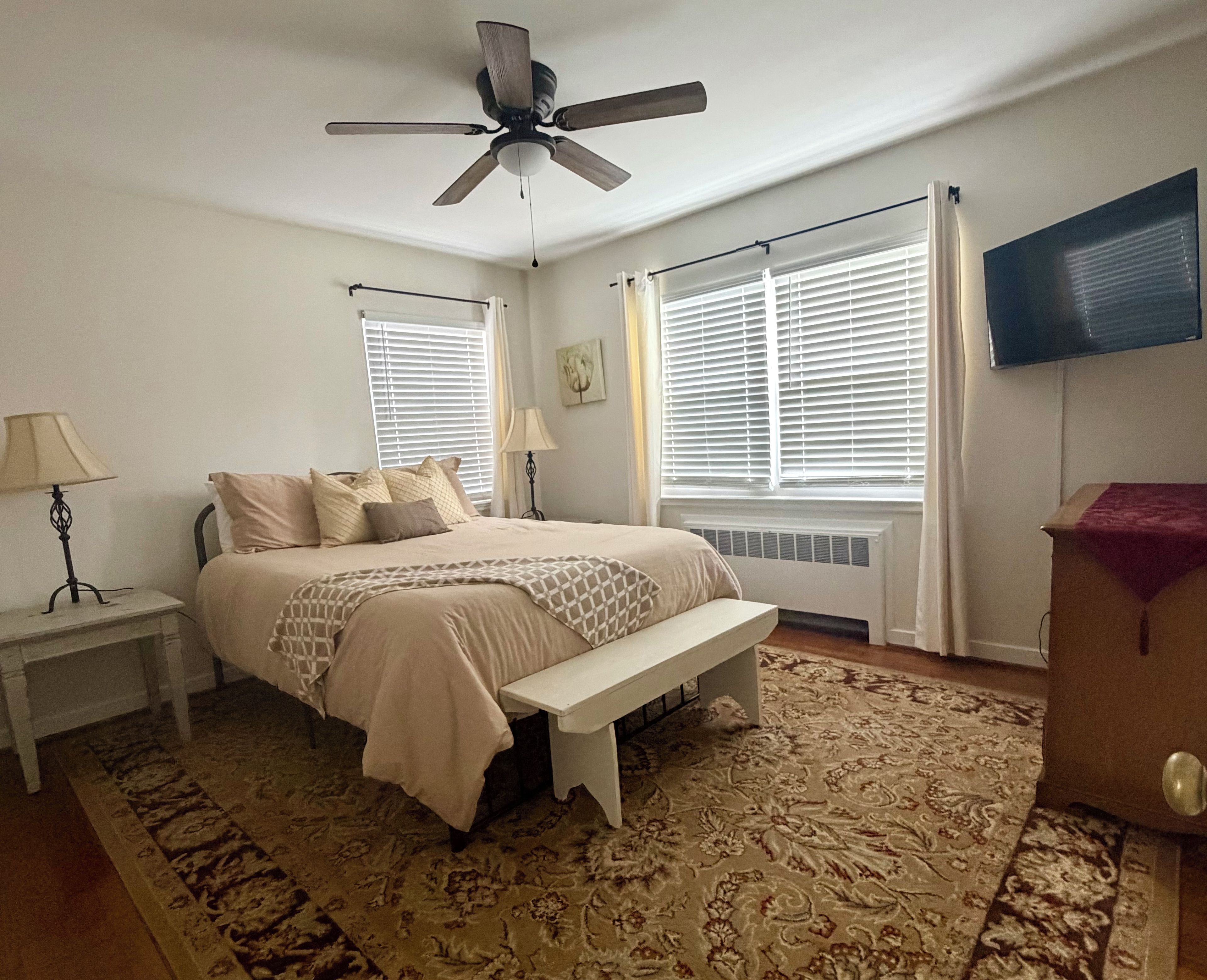 A bedroom with a bed, bedside tables, a ceiling fan, a radiator, and a window with blinds.
