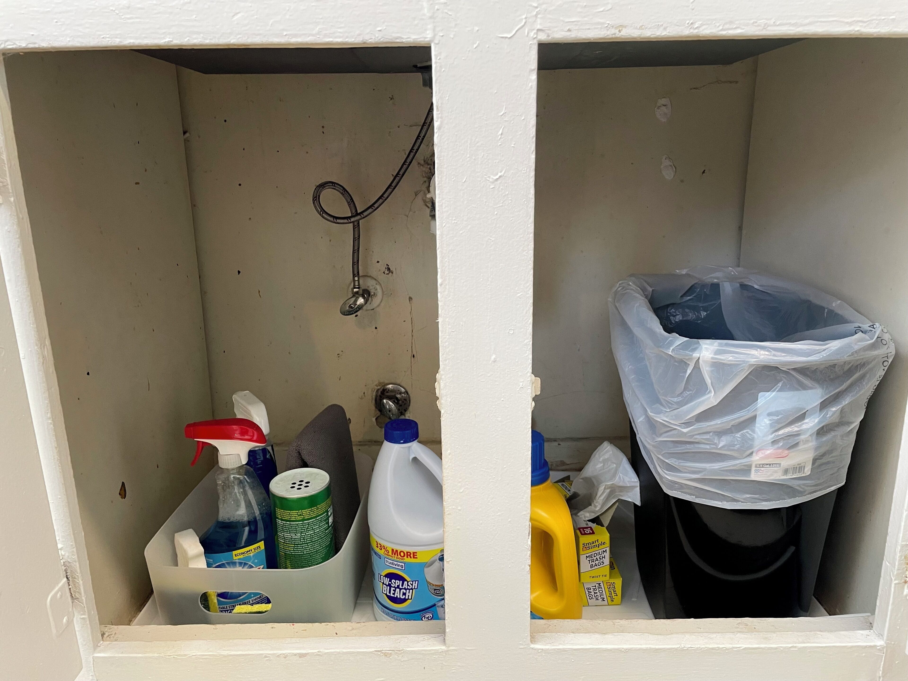 A storage area with cleaning supplies, including a bottle of bleach, a spray bottle, and a trash bag.