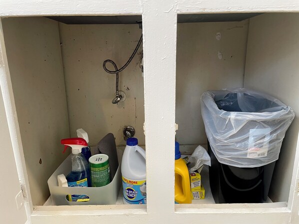 Zona de almacenamiento con productos de limpieza, incluyendo una botella de lejía, un pulverizador y una bolsa de basura.