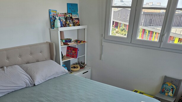 Une chambre à coucher avec un lit, une étagère blanche contenant des livres et des jouets pour enfants, et une fenêtre donnant sur des immeubles.