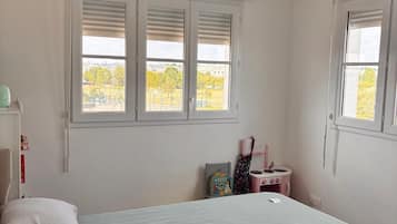 Un dormitorio con una cama, dos ventanas, una mesita con una silla rosa y un estante en la pared.