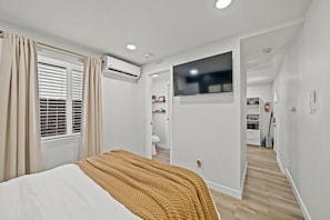 A bedroom with a bed, an air conditioning unit, a window with curtains, and a view into a bathroom and kitchen area. - La Esperanza Unit B Cozy Central SD Retreat w/ A/C (National City)