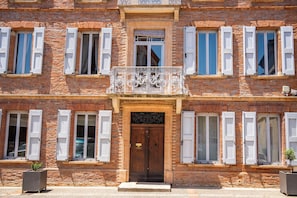 Un edificio de dos pisos con una entrada central, contraventanas blancas y un balcón decorativo.