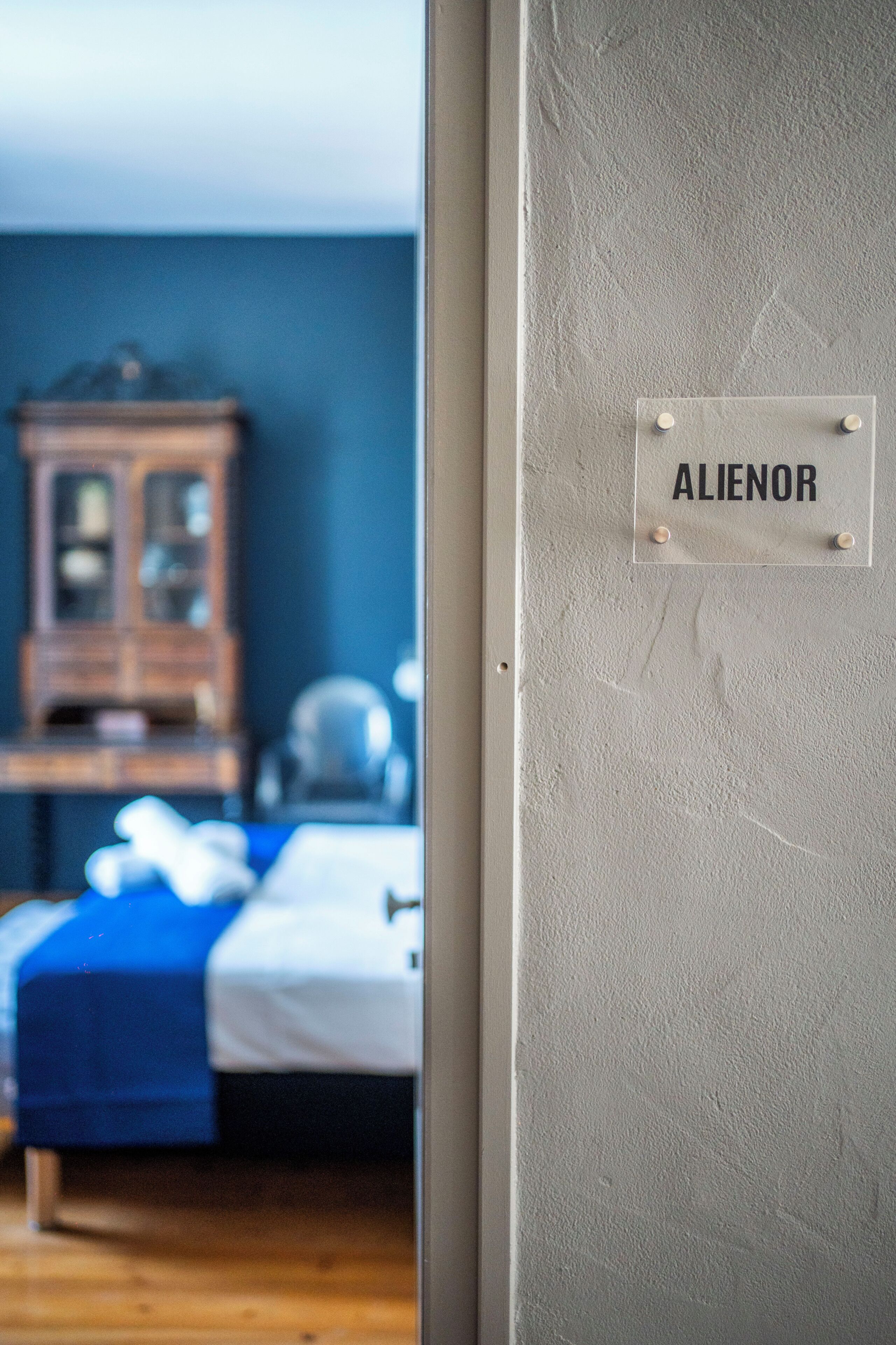 A room with a bed, a wooden cabinet, and a wall-mounted sign reading 'ALIENOR'.