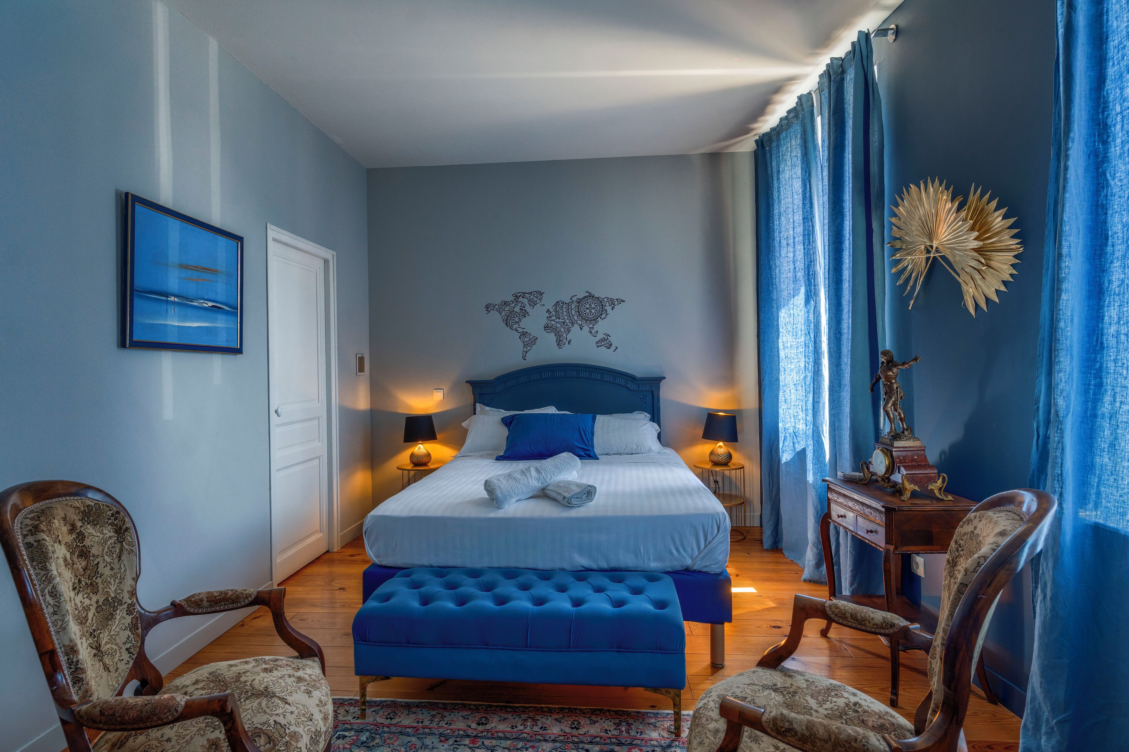 A bedroom with a large bed, two nightstands, a blue upholstered bench, and a wooden chair.