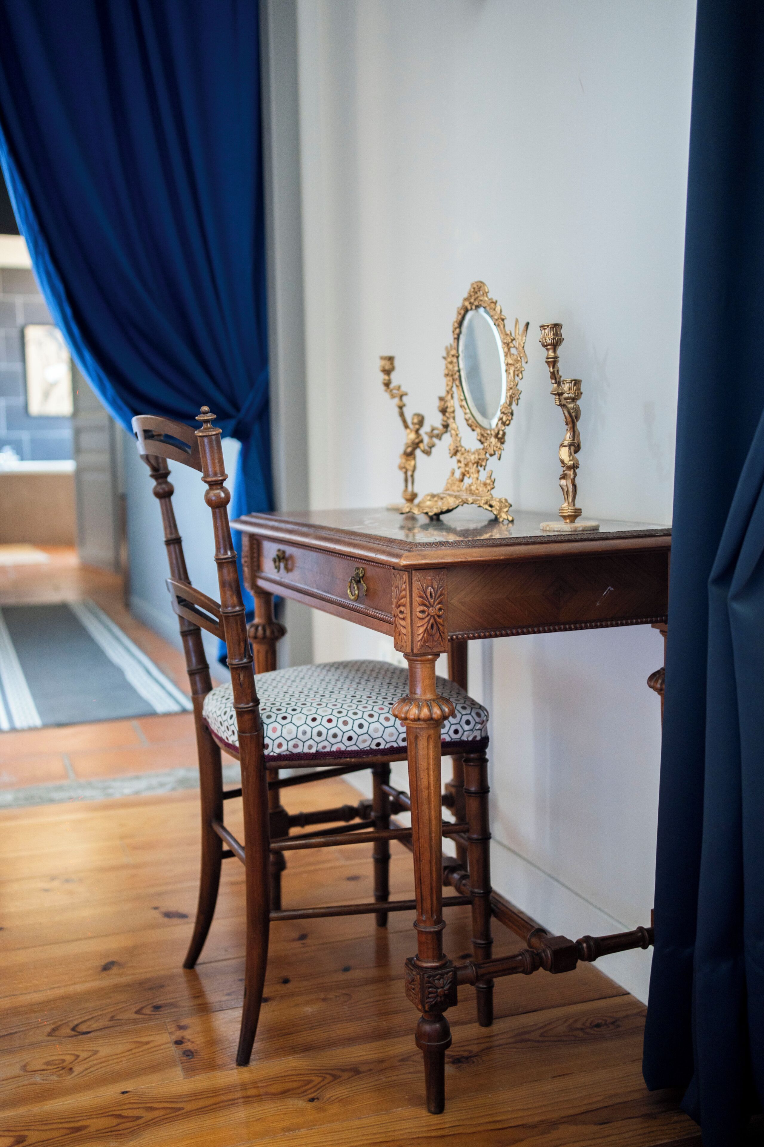 A wooden dining table with a mirror and candle holders, a chair with a patterned cushion, and a blue curtain partially drawn.