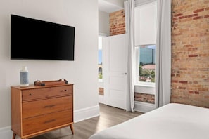 Ein Schlafzimmer mit Bett, Kommode, flachbildfernseher an der Wand und einem Fenster mit Vorhängen.