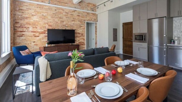 A modern living room with a brick wall, a flat-screen TV, a sofa, and a dining table set for four. - Stunning 1BR Walkable Loft above Palafox (Pensacola)