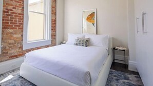 A bedroom with a brick wall, a large window, a bed with white bedding, a nightstand, and a framed picture on the wall. - Stunning 1BR Walkable Loft above Palafox (Pensacola)