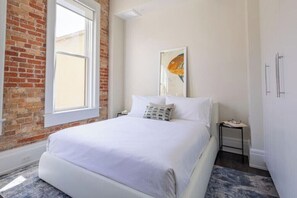 Ein Schlafzimmer mit Backsteinwand, großem Fenster, einem Bett mit weißer Bettwäsche, einem Nachttisch und einem gerahmten Bild an der Wand.