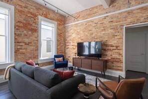 A living room with a brick wall, a sofa, a blue chair, a television, and a coffee table. - Stunning 1BR Walkable Loft above Palafox (Pensacola)