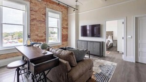 A modern living room with a brick wall, a large window, a flat-screen TV, and a cozy seating area. - The Brent Lofts A Quiet Place 303 (Pensacola)