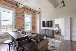 A modern living room with a brick wall, a large window, a flat-screen TV, and a cozy seating area. - The Brent Lofts A Quiet Place 303 (Pensacola)