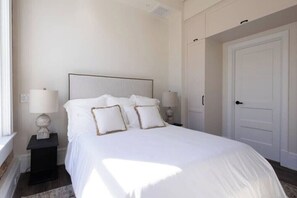A neatly made bed with white bedding and two bedside lamps, a closed door, and a window on the left. - Historic Charm Southern Exposure (Pensacola)