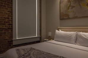 A hotel room with a bed, a nightstand, a lamp, a window with blinds, and a wall with a painting.